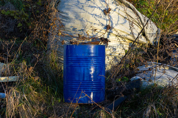A blue barrel is surrounded by dry grass and overgrown brush in a rural location, with discarded...