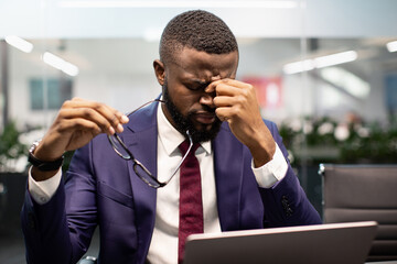 Closeup of exhausted black man manager or businessman sitting at workdesk in front of laptop at modern office, holding glasses and rubbing nose bridge, having headache, burnout at work concept
