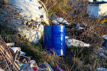 A blue barrel sits among piles of trash and overgrown plants in a desolate outdoor area under...