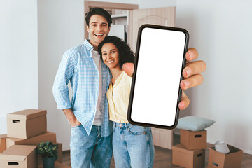 A happy couple stands in a room filled with moving boxes. They hold a smartphone with a blank screen, smiling as they enjoy their fresh start in a new home.