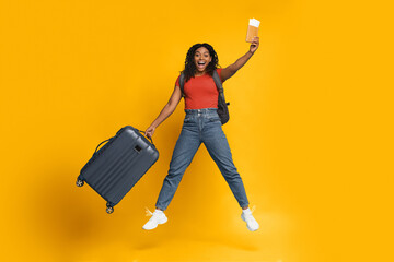 A joyful woman is jumping in the air, holding a suitcase in one hand and travel snacks in the other. She smiles brightly, wearing casual clothes with a backpack on her back.