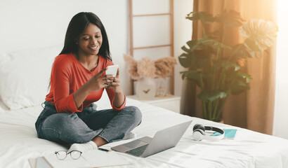A young woman sits comfortably on her bed, smiling as she uses her smartphone. A laptop rests nearby, alongside headphones and a notebook, creating a relaxed study atmosphere.