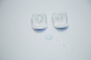 Close-up of one-day contact lenses in disposable protective packaging lying in the center on a white background with a transparent contact lens next to it, surrounded by water droplets. The concept of