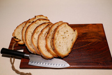 Fresh slices of rustic bread rest elegantly on a dark wooden board, complemented by a chef’s knife that adds a touch of authenticity and home-cooking charm.
