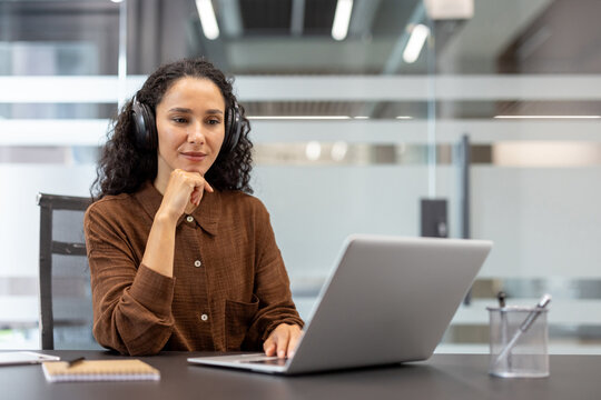 Young woman with curly hair wearing headphones and an earth tone shirt. Concentrating while using a laptop from a desk in a contemporary co-working office. Focusing on an online webinar or remote work - Powered by Adobe