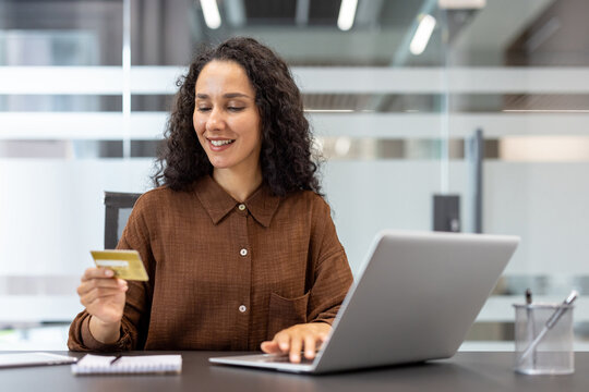 Woman smiling and holding a gold credit card while performing an online transaction on a laptop, representing digital commerce and secure internet banking in an office setting
