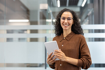 Confident businesswoman with curly hair and glasses, smiling while holding a digital tablet in a...