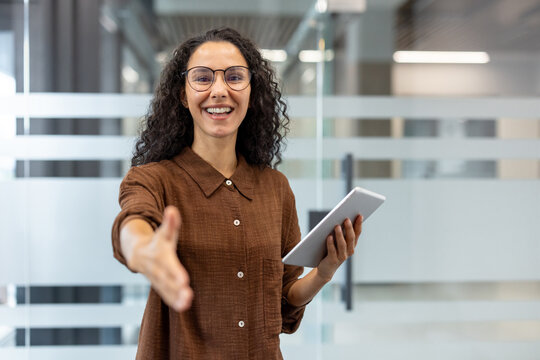 Professional businesswoman in a modern office smiling and reaching out for a handshake while holding a tablet, conveying welcome, partnership, hiring and confident collaboration