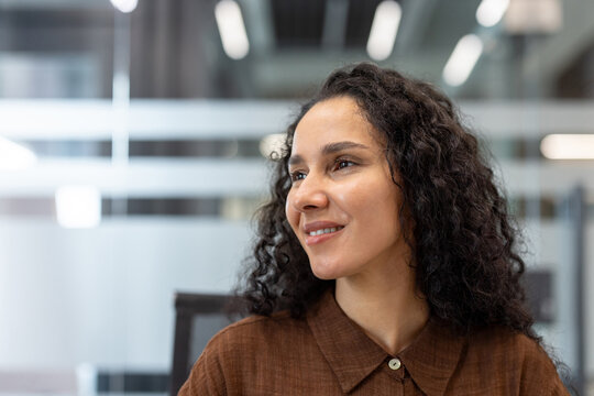 Young businesswoman with curly hair smiling expressing hope and positivity, looking to the side confidently in a contemporary open-plan office setting