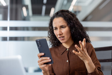 Young businesswoman with curly hair expressing anger and frustration while reading a message on her mobile phone, experiencing problems or bad news at her modern workplace