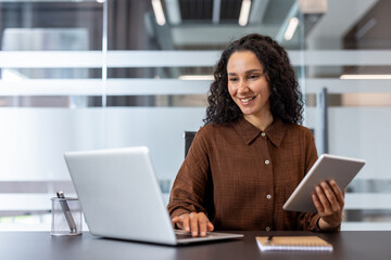 Young businesswoman with curly hair smiling and actively engaged in work, balancing a laptop and a tablet at her modern office desk, showcasing efficiency and productivity