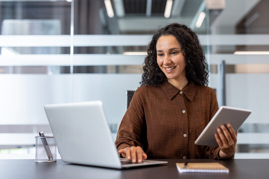 Young businesswoman with curly hair smiling and actively engaged in work, balancing a laptop and a tablet at her modern office desk, showcasing efficiency and productivity