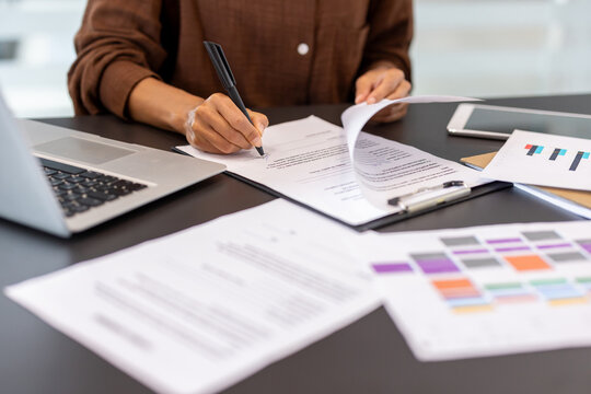 Person signing a legal business contract or agreement document with a pen, sitting at an office desk with other paperwork, laptop, and charts during work