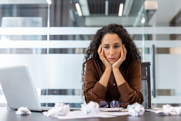 Businesswoman feeling overwhelmed and frustrated while working at an office desk, surrounded by crumpled papers, symbolizing stress, exhaustion, and creative block