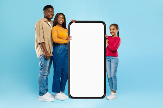 A cheerful Black family stands in front of a blue wall, leaning on a large smartphone with a blank screen. The mom, dad, and daughter display excitement while recommending a new app to viewers.