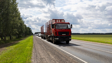 Logging truck with a loader drives along the road. A front view of a long, empty red industrial logging trailer moving along the highway against a blue summer sky.