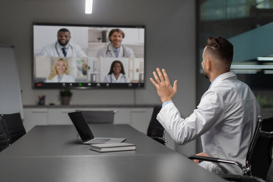 A middle eastern doctor participates in a video call with international colleagues. He waves while viewing a digital display showing faces of other healthcare professionals.
