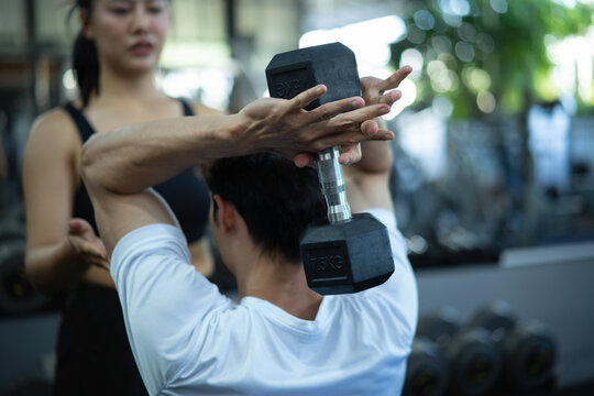 A woman assists and checks a man's dumbbell lifting form during a workout at the gym - Powered by Adobe