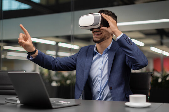 Excited businessman in a modern office sits at his desk wearing VR glasses. He engages with virtual content, pointing at something with enthusiasm while connected to advanced technology.