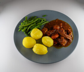 A hearty beef stew is served with tender boiled potatoes and fresh green beans on a blue plate. The meal is prepared at home in a cozy kitchen setting, creating a warm atmosphere