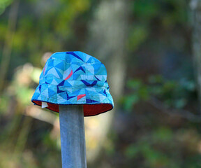 A vibrant bucket hat featuring geometric patterns is placed on a wooden post surrounded by greenery. Sunlight filters through the trees, creating a serene atmosphere