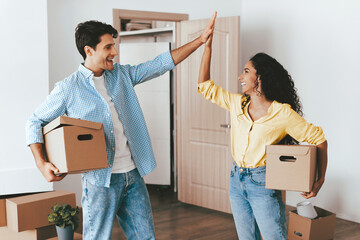 A happy couple shares a high five while surrounded by cardboard boxes in their new apartment. They look excited and ready to start their new chapter together.