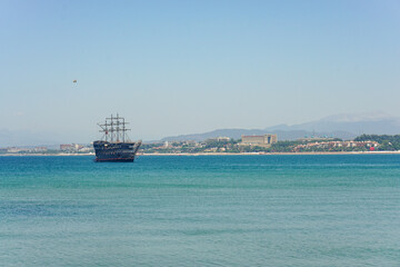 A large, old ship with masts sails along the coast near the town of Side in Turkey It's hot and hazy, and the silhouette of the mountains is visible in the background