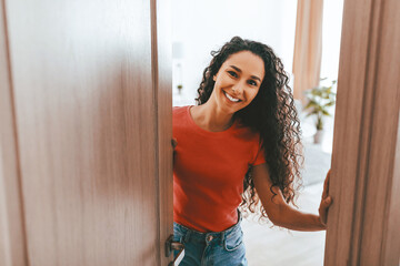 A woman with curly hair stands at an open door, smiling joyfully. Sunlight fills the room, enhancing the inviting atmosphere. Her casual outfit suggests comfort and ease.