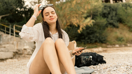 Woman sitting on a pebble beach, using her smartphone and enjoying a beautiful sunny day. Relaxing vacation moment by the sea, leisure and technology concept.