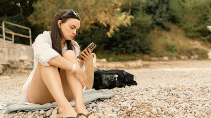 Woman sitting on a pebble beach, using her smartphone and enjoying a beautiful sunny day. Relaxing vacation moment by the sea, leisure and technology concept.