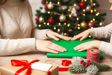 Two hands preparing green wrapping paper for a gift on a wooden table, surrounded by festive decorations, presents, and a Christmas tree, capturing the holiday spirit and joy of gift-giving