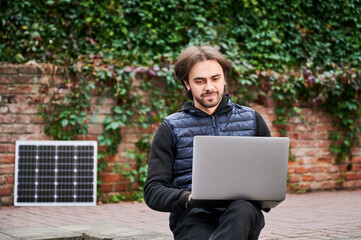 Man sits outdoors, working on laptop against backdrop of ivy-covered brick walls. Solar panel rests nearby, highlighting sustainable, eco-friendly workspace in serene, natural urban environment.