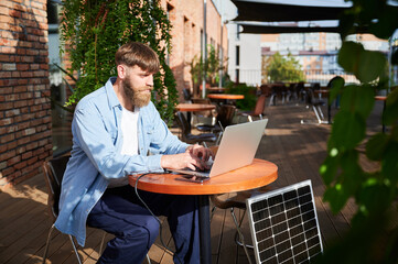 Man works on laptop and smartphone at outdoor table. Solar panel nearby, emphasizing sustainable, modern eco-friendly workspace that combines technology and sustainability.