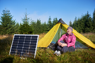 Happy woman sits in front of yellow tent with solar panel, enjoying sunny morning in grassy field. Concept of eco-friendly camping, renewable energy. On background lush green trees and clear blue sky.