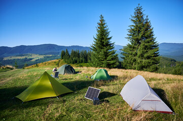 Tourist tents with solar panel and portable power station nearby in grassy field. Scene surrounded by rolling hills, tall pine trees under clear blue sky, showcasing eco-friendly camping setup.