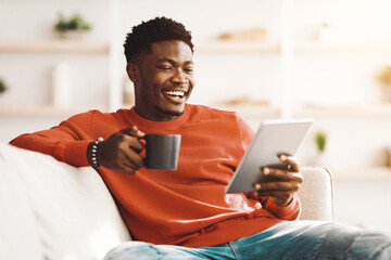 A young man in a red sweater sits comfortably on a couch, smiling as he reads on his tablet and holds a cup of coffee in a bright and inviting living room setting.