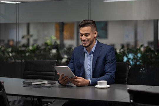 Handsome young Middle Eastern entrepreneur sits at a sleek table in a modern office. He smiles while using a digital tablet to manage his business with a mobile application during daylight.