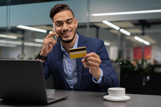 Young businessman is smiling while holding a credit card in one hand and speaking on a mobile phone. He appears satisfied after making an online purchase in a modern office setting. - Powered by Adobe