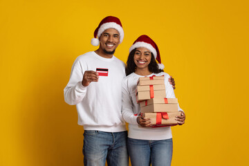 Two excited individuals wear Santa hats and hold a credit card and wrapped presents. They stand against a bright yellow backdrop, embodying the festive spirit of the holiday season.