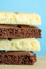 Stack of aerated white and milk chocolate bars on a wooden surface against a light blue background. The close-up highlights the bubbly texture and contrasting colors, creating a clean, minimal, and ap