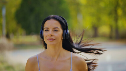 Young woman jogging through a green park in sportswear and wireless headphones, smiling as she enjoys a morning run, fitness, fresh air and focused, energetic movement