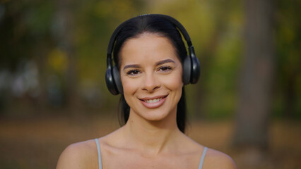Brunette woman wearing wireless headphones, smiling at camera while relaxing in a park, enjoying...