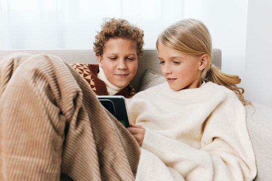 Two children sit close under a warm blanket on a sofa, sharing a tablet and smiling as they explore a digital story or game together.
