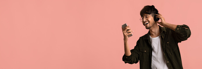 A joyful young man listens to music on his headphones while holding a smartphone in one hand. He is smiling and appears to be dancing against a pink background.