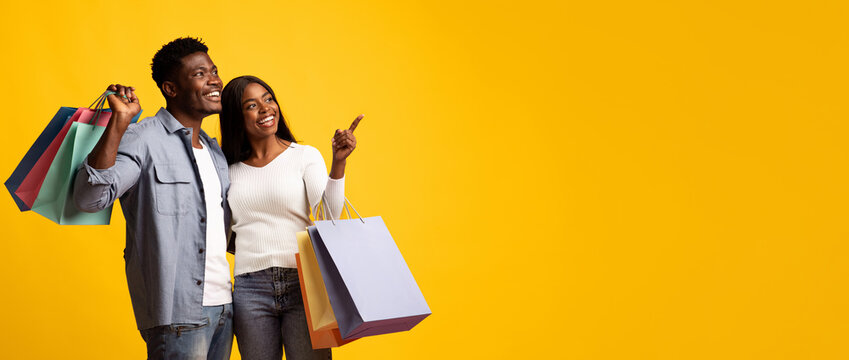 Two cheerful individuals stand together holding shopping bags. The couple appears to be having a good time, smiling and pointing at something off-screen. The vibrant yellow backdrop adds energy.