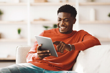A young man sits comfortably on a couch in a cozy living room, smiling and engaging with a tablet. Natural light fills the space, creating a warm atmosphere.