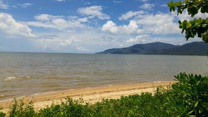 Cairns, Australia - 5 March 2018: Coastal Sandy Beach with Vegetation, Calm Sea, and Mountainous Horizon.  The sky is partly cloudy with patches of blue, enhancing the relaxed tropical atmosphere. 