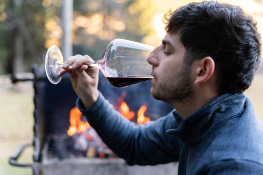 Young man drinking red wine by barbecue fire outdoors