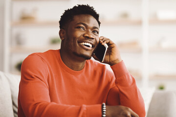 A man wearing a red sweater enjoys a cheerful conversation on his smartphone while seated in a comfortable indoor space. His smile reflects happiness and engagement with the call.