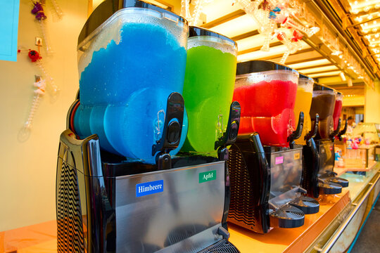 A colorful row of slush machines filled with bright blue, green, red, yellow, and dark frozen drinks, labeled with different flavors and displayed at a lively refreshment stand.